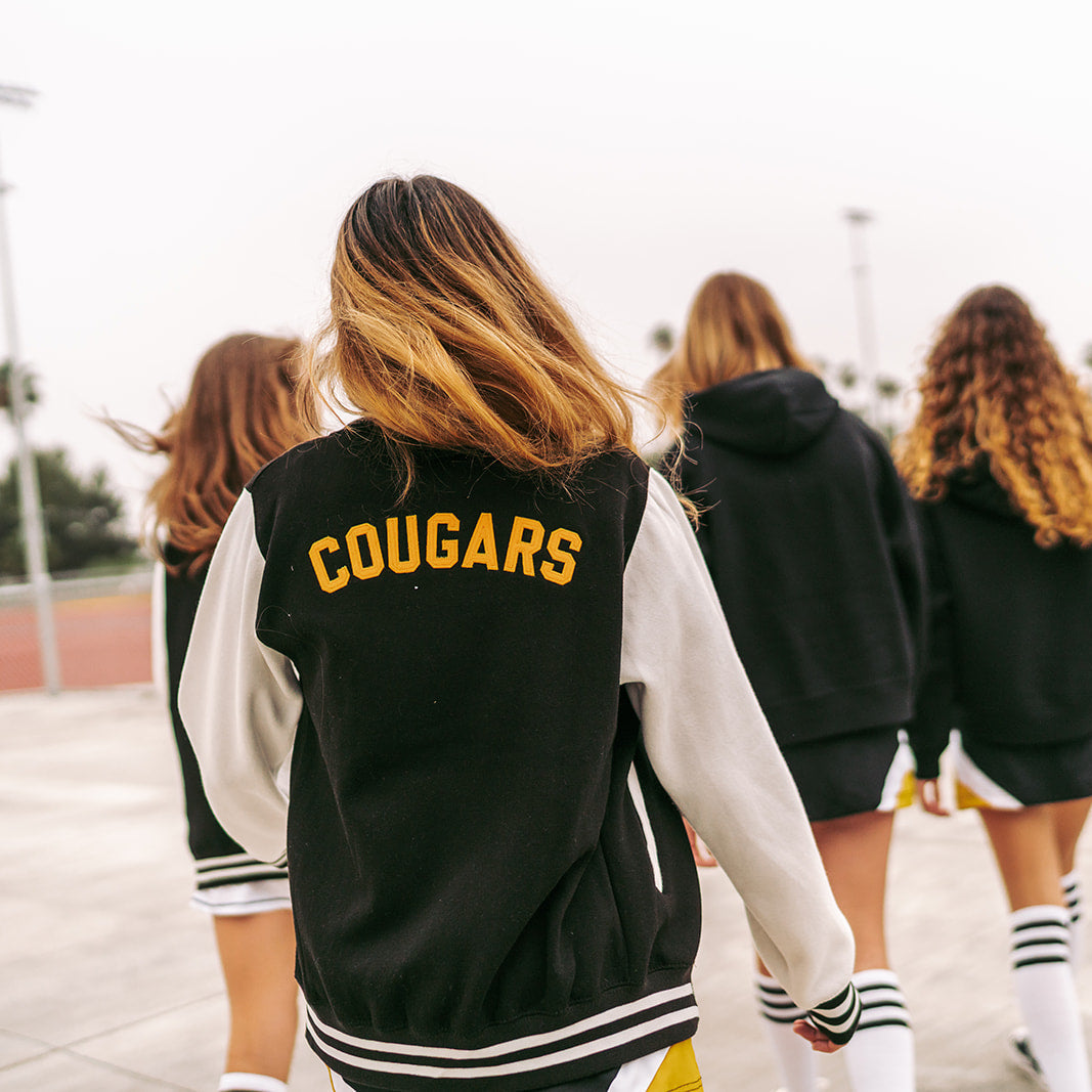 Four girls wearing 'COUGARS' letterman jackets walking together outdoors.
