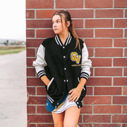 Young woman wearing a black and white varsity jacket with a logo, standing against a brick wall.